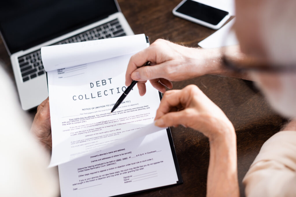 Close-up of hands signing a debt collection form on a desk, with a laptop and smartphone nearby.